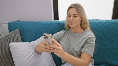 Modern bir oturma odasındaki deniz mavisi koltukta akıllı telefon kullanan beyaz bir kadın.