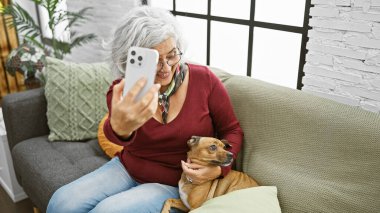 Beyaz saçlı kadın evde selfie çekerken köpekle eğleniyor.