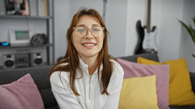 Gözlük takan neşeli genç bir kadın, renkli yastıklarla çevrili evinde rahatça oturuyor..