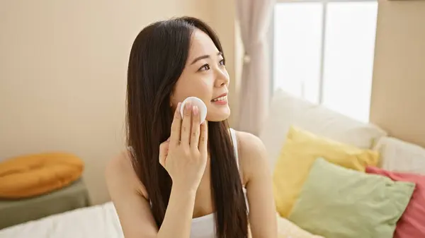 stock image A young asian woman applies makeup in a bright, cozy bedroom setting, embodying beauty and routine.