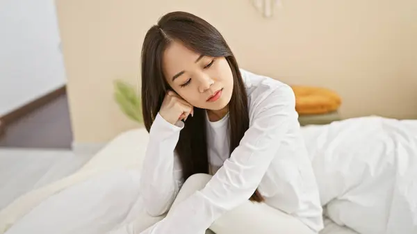 stock image Pensive asian woman in a white shirt sitting alone in a bright bedroom reflecting on her thoughts.