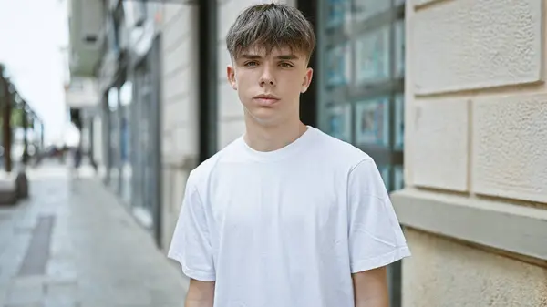 stock image A young caucasian male teenager with blond hair in a white shirt stands on an urban street, portraying a casual outdoor setting.