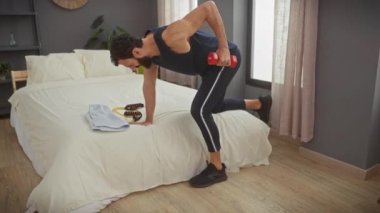 Atletik giysili sakallı bir adam yatak odasındaki bir dambılla tek bacaklı bir kürek çeker..