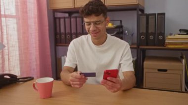 Ofisteki genç İspanyol bir adam akıllı telefon kullanıyor ve kredi kartı tutuyor. Elinde dosyalar ve kahve fincanı olan bir masada oturuyor. İlginç bir iş yeri ya da online alışveriş merkezi yaratıyor..