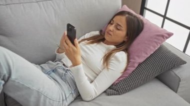 Genç ve çekici bir kadın evdeki bir kanepede uzanmış, rahat bir tavırla akıllı telefonuna bakıyor, modern ve rahat bir oturma odası hazırlıyor..