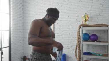 Spor salonundaki yakışıklı zenci bir adam boynunda havluyla geziyor. Spor aletleri ve spor eşyalarıyla çevrili..