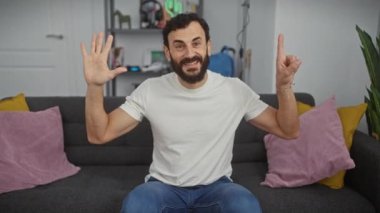 Neşeli orta yaşlı bir adam, altı numaranın neşeli bir hareketini parmaklarıyla gösteriyor, güvenli bir şekilde oturma odasını işaret ediyor, beyaz bir tişört giymiş..