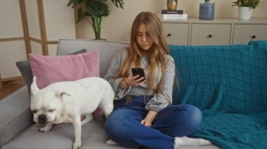 Beyaz köpek köpeği yanında renkli yastıklarla süslenmiş gri bir kanepede dururken, genç sarışın beyaz bir kadın oturma odasında cep telefonuyla hayal kırıklığına uğramış bir şekilde oturuyor..