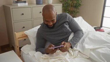 Yatakta yatan Afrikalı genç adam rahat bir yatak odasında şifoniyer ve bitkilerle telefon kullanıyor.
