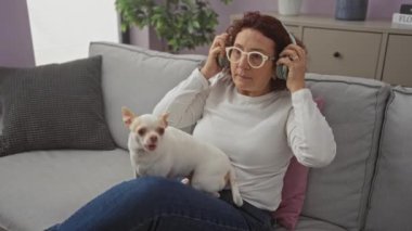 Oturma odasında elinde Chihuahua köpeği olan, cep telefonu kullanan ve kulaklık takan olgun bir beyaz kadın..