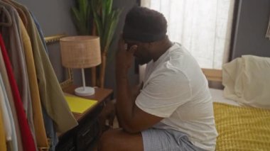 Sakallı Afrikalı genç bir Amerikalı yatak odasında bir yatağa oturmuş, günlük kıyafetlerini giymiş ve düşünceli görünüyor, giysileri yakında asılı ve komodinin üzerinde bir lamba var..