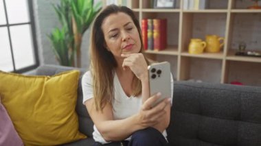 Düşünceli İspanyol kadın oturma odasında elinde akıllı telefon, mesajlaşmayı düşünüyor.