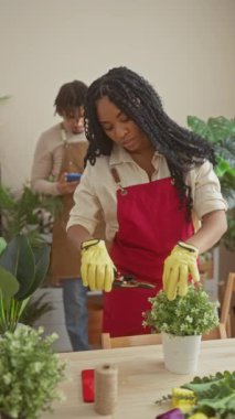 Kırmızı önlüklü bir kadın çiçekçi, arka planda telefonu olan bir adam dururken içeride çiçek düzenliyor..