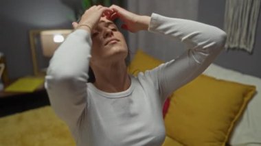 Genç bir kadın, sarı yastıklı ve loş ışıklandırmalı rahat bir yatak odasında saçını düzeltiyor..