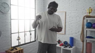 Sakallı Afrikalı genç bir Amerikalı spor aletleri ve duvardaki saatle dolu bir spor salonunda kulaklık takarken telefonuna bakıyor..