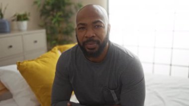 Yatak odasında, sarı yastıklı bir yatakta oturan, gri gömlekli, arka planda bitkilerle süslenmiş bir oda ve çekmeceli bir çekmece.