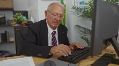 Ofisin içinde bilgisayar ve masa başında çalışan yaşlı beyaz bir adam, kitapların ve bitkilerin arka planıyla monitöre doğru hareket ederken duygularını ifade ediyor..