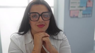 Ofis ortamında gülümseyen İspanyol kadın, gözlüklü ve beyaz gömlekli, arka planı gri duvarlı ve hafif ofis dekorasyonlu.