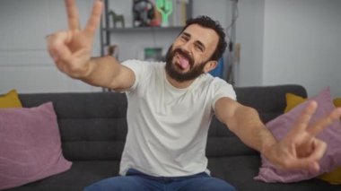 Neşeli orta yaşlı bir adam zafer işareti gösteriyor. İki eli, dili açık, evinde beyaz tişört giyiyor..