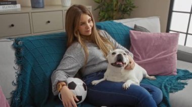 Genç sarışın bir kadın, köpeği ve futbol topuyla rahat bir oturma odasında rahatlıyor..