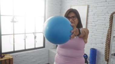 Spor salonunda orta yaşlı bir İspanyol kadın mavi bir egzersiz topu tutuyor. Spor salonunda spor ve spor faaliyetleri sergiliyor..