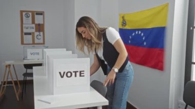 Venezuela seçimlerinde oy veren çekici genç bir kadın seçim odasında arka planda Venezuela bayrağı olan kulübelerin yanında duruyor..