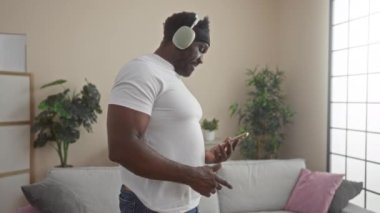 Sakallı, genç bir Afro-Amerikan adam, oturma odasında şık bir şekilde bitkiler ve yumuşak mobilyalarla süslenmiş bir şekilde telefonunu kullanırken kulaklıkla müzik dinliyor..