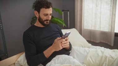 Siyah tişörtlü, orta yaşlı sakallı bir adam yatak odasında rahatlar ve içerik ifadesiyle akıllı telefonunu karıştırır..