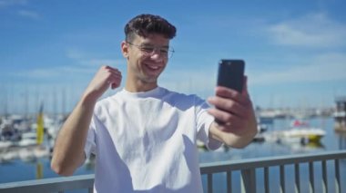 Genç İspanyol bir adam güneşli bir günde deniz kenarındaki bir liman ortamında telefon görüşmesi yapıyor. Bir eliyle akıllı telefonu tutarken elini yumruk atarak sevinçten havalara uçuyor..