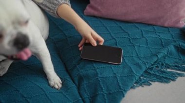 Genç bir kadın mavi bir kanepede, yanında mutlu beyaz köpek arkadaşıyla birlikte, elinde akıllı bir telefon tutarak rahatlıyor..