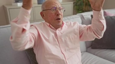 Gözlüklü yaşlı adam, sıcak bir oturma odasında kanepede otururken heyecandan kollarını kaldırıyor..