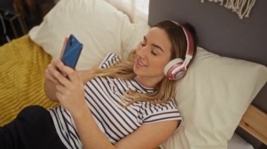 Sarı saçlı genç bir kadın yatak odasında uzanmış, akıllı telefonunu kullanıyor ve kulaklık takıyor..