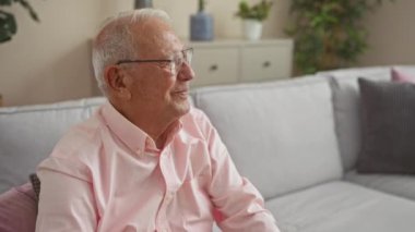 Pembe tişörtlü, mutlu, kır saçlı yaşlı bir adam rahat bir şekilde otlarla dolu modern bir oturma odasında oturur..