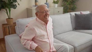 Pembe tişörtlü, gri saçlı yaşlı bir adam oturma odasında yeşil bir kanepede otururken acı içinde kıvranıyor. Etrafı bitkiler ve modern dekorlarla çevrili..