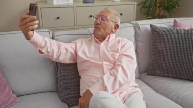 Pembe tişörtlü, olgun, gri saçlı bir adam modern oturma odasındaki rahat gri bir koltukta otururken akıllı telefonuyla video görüşmesi yapıyor..