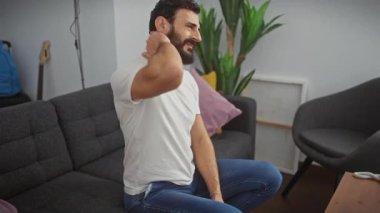 Evde acı çeken orta yaşlı bir adam, beyaz tişört giyiyor ve kaslı bölgeye dokunuyor, evde sağlık sorunları yaşıyor.