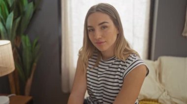 Güzel sarışın kadın yatak odasında bir yatakta oturuyor. Arkasında çizgili gömlek ve bitkiler var..