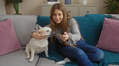 Sarı saçlı genç bir kadın oturma odasında oturuyor, küçük köpeğiyle eğleniyor ve elinde uzaktan kumandayla....