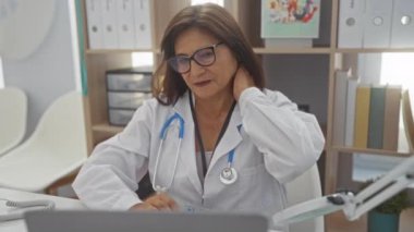 Beyaz önlüklü ve steteskoplu orta yaşlı bir İspanyol kadın, boyun ağrısı çekiyor, sağlık ekipmanları, kitaplar ve klasörlerle çevrili bir klinikte oturuyor..