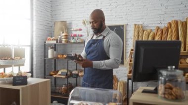 Fırında sakallı yakışıklı kel bir adam tablet kullanıyordu. Etrafı çeşitli ekmek ve hamur işleriyle çevrili mavi önlük giyiyordu.