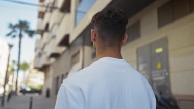 Latin kökenli genç bir adam şehir caddesinde gün ışığında yürür, binalar ve palmiye ağaçlarıyla dolu bir şehir ortamını gösterir..