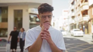 Gündüz vakti kalabalık bir caddede öksüren genç İspanyol adam kendini kötü hissediyor ve göğsüne dokunuyor..