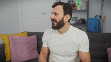 Öfkeli orta yaşlı bir adam evde kızgın bir sesle buhar çıkarıyor, elleri klasik beyaz bir tişört giyerken öfkesini ağzının üstüne çıkarıyor.