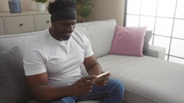 Afro-Amerikalı bir adam oturma odasındaki bir kanepede akıllı telefon kullanarak elini kaldırarak kutlama yapıyor..