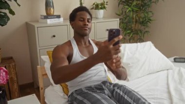 Genç adam yatak odasında akıllı telefonuyla selfie çekiyor. Kolsuz tişört ve pijama giyiyor. Etrafı modern dekorlarla çevrili.