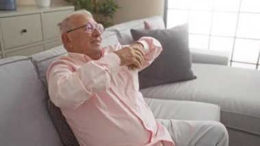 Yaşlı beyaz bir adam evinde şık bir oturma odasında rahatça dinleniyor. Dairesinde rahat bir kanepede otururken kollarını uzatıyor..