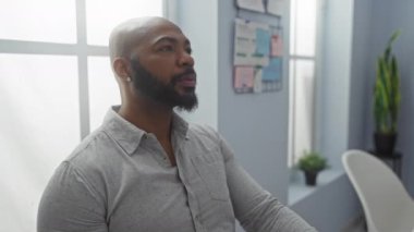 Yakışıklı, kel, sakallı, gri gömlekli, modern ofiste oturan, arkasında bitki ve pencere olan bir adam.