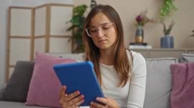 Video izlerken mavi tablet kullanan genç bir kadın oturma odasında yastıklar, bitkiler ve kitaplıklarla konuşuyor..