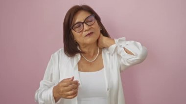 Orta yaşlı, kısa saçlı ve gözlüklü bir İspanyol kadın, pembe arka planda beyaz giyinmiş, rahatsız bir şekilde boynuna dokunuyor..