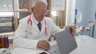 Hastane odasında dizüstü bilgisayarda çalışan kıdemli erkek doktor. Arka planda tıbbi malzemeler ve kitaplıklar var..